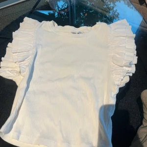 Vit topp med volangärmar  - Supersöt vit ribbad topp från Zara med korta ärmar och stora volangdetaljer. Mjuk och stretchig, passar till många olika outfits. Ganska liten i storlek men annars jättesöt!🩷 Tveka inte att skriva till mig för ytterligare frågor!🫶😊🩷