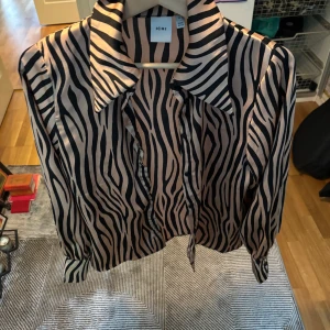 Zebramönstrad blus med knyt från ICHI - Säljer en snygg zebramönstrad blus från ICHI i beige och svart. Blusen har lång ärm, klassisk krage och knytband vid halsen. Materialet är glansigt och känns lyxigt, perfekt för att sticka ut lite extra.