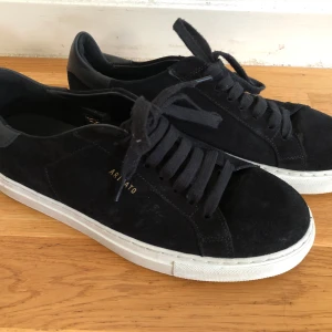 Svarta sneakers från Axel Arigato 40 - Stilrena svarta sneakers från Axel Arigato med ovandel i mocka och detaljer i skinn. Skorna har snörning, vit platt sula och diskret logga i guld på sidan. Perfekta för en clean och modern look. Storlek 40, fint skick!