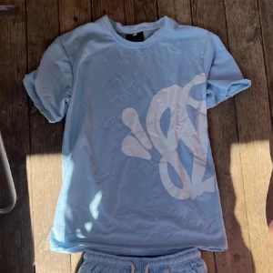 Ljusblå t-shirt från Syna world - Säljer en ljusblå t-shirt från Syna world med ett stort vitt grafiskt tryck på både fram- och baksidan. T-shirten har rund hals och korta ärmar. Perfekt för dig som gillar streetwear och vill sticka ut med en unik design.