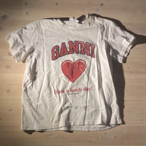 Vit t-shirt med tryck från Ganni - Vit t-shirt från Ganni med rött hjärta och texten 'Have a lovely day!' framtill. Klassisk passform med rund hals och korta ärmar. Perfekt statement-plagg för en cool och avslappnad stil.