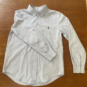 Ljusblå skjorta från Ralph Lauren - Klassisk ljusblå skjorta från Ralph Lauren med broderad logga på bröstet. Skjortan har lång ärm, knappar framtill och krage. Perfekt för en stilren och tidlös look.