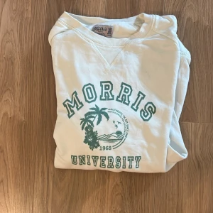 Vit sweatshirt från Morris University - Vit sweatshirt från Morris med grönt tryck framtill där det står 'Morris University' och ett palm-motiv. Klassisk rund halsringning och mjukt material, perfekt för en avslappnad stil.