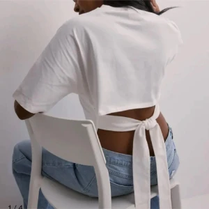 Vit t-shirt med knyt i ryggen - Säljer en vit croppad t-shirt med korta ärmar och snygg knytning i ryggen. Perfekt till jeans eller kjol för en trendig look. Enkel och stilren design som passar till många outfits.