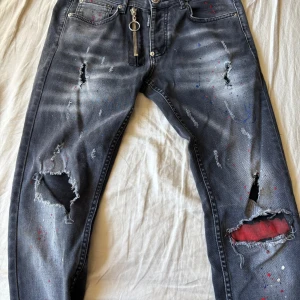 Svarta distressed jeans från DSQUARED2 - Säljer ett par svarta jeans från DSQUARED2 med slitna detaljer, hål och färgstänk. Byxorna har dragkedja med ring, klassisk femficksdesign och röd patch på bakfickan. Snygg streetstil med unik look.