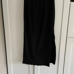 Svart kjol med slits från Holys - Säljer en svart kjol från Holys. Klänningen har en stilren design med en slits på sidan som ger en snygg detalj. Perfekt för dig som gillar minimalistisk stil och vill ha något enkelt men elegant.
