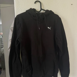 Svart hoodiejacka från Puma - Svart hoodiejacka från Puma med dragkedja framtill och huva med dragsko. Klassisk design med Puma-logga på bröstet. Perfekt för vardagsbruk eller till träning. Mjuk och bekväm med fickor på sidorna. Snarare S i storleken! 