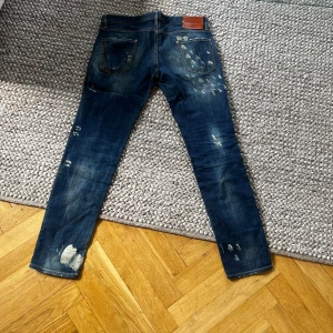Mörkblå jeans från Dsquared2 - Säljer ett par mörkblå jeans från Dsquared2 med slitna detaljer och unika blekta partier. Klassisk femficksmodell med raka ben och läderpatch bak i midjan. Perfekta för dig som gillar en edgy och trendig look.