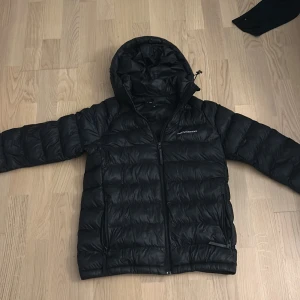 Svart dunjacka från Peak Performance - Säljer en svart dunjacka från Peak Performance med huva och dragkedja framtill. Jackan har två sidofickor med dragkedja och är perfekt för kyligare dagar. Klassisk och stilren design med diskret logga på bröstet.