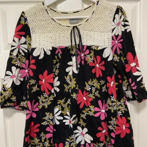 Somrig blus/tunika med fina detaljer  - Nyskick! Säljer en somrig blus/tunika från Lindex med stora blommor i vitt, rött och rosa samt gröna blad. Ovanpartiet är virkat i vitt och har en knytning vid halsen. Blusen har trekvartsärm och en lös passform. Jättefin och ovanlig!