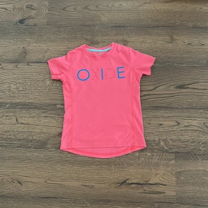 Rosa t-shirt från Oxide - Rosa t-shirt från Oxide med blå och rosa tryck framtill där det står 'OIDE'. T-shirten har rund halsringning med turkos kant och är kortärmad. Perfekt för en färgglad och sportig look.