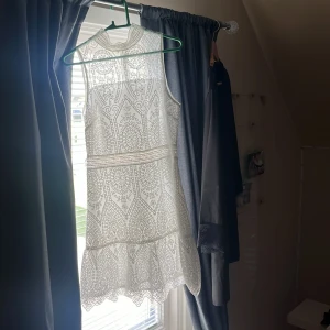 Vit spetsklänning med volang - Superfin vit ärmlös klänning med spetsdetaljer och volang nertill. Klänningen har hög hals och ett vackert mönster i spetsen som ger en romantisk känsla. Perfekt för sommarens tillfällen eller studentfirande.
