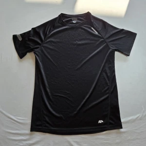 Svart sport t-shirt från Karhu - Svart sport t-shirt från Karhu i syntetmaterial. T-shirten har korta ärmar och en klassisk rund halsringning. Perfekt för träning eller sportaktiviteter. Diskret logga på bröstet och axeln.
