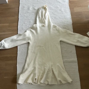 Vit stickad klänning med volang från Zara - Mysig vit stickad klänning från Zara med huva och långa ärmar. Klänningen har en volang nertill som ger en söt touch. Perfekt för kyligare dagar och enkel att styla med tights eller leggings.