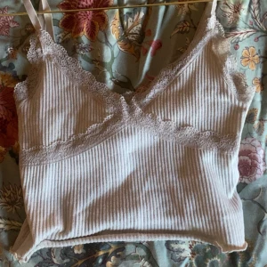 Vit ribbad linnetopp med spetsdetaljer - Säljer en söt vit ribbad linnetopp med tunna axelband och spets längs urringningen och under bysten. Perfekt att styla till jeans eller kjol för en trendig look.