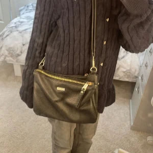 Svart axelväska från Don Donna - Säljer en svart axelväska från Don Donna i strukturerat skinnimitation. Väskan har två stora fack med guldfärgade dragkedjor och justerbar axelrem. Perfekt storlek för det viktigaste och stilren design med metallogo framtill.