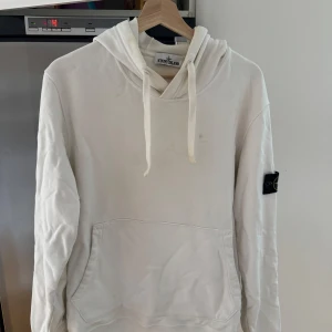 Vit hoodie från Stone Island - Säljer en stilren vit hoodie från Stone Island med klassisk huva och känguruficka. Tröjan har den ikoniska Stone Island-loggan på vänster ärm och dragsko i huvan. Perfekt för dig som gillar streetwear och enkel design. Köpt på Nk
