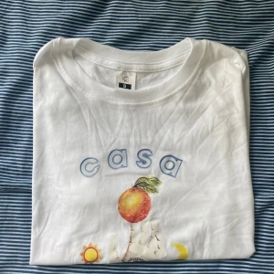 Vit t-shirt med tryck från Casablanca  - Vit t-shirt med färgglatt tryck framtill som visar en hand som håller ett äpple, med sol, moln och måne runtom. Texten 'casa sport' i blått. Klassisk passform och rund halsringning. Perfekt för en avslappnad stil.