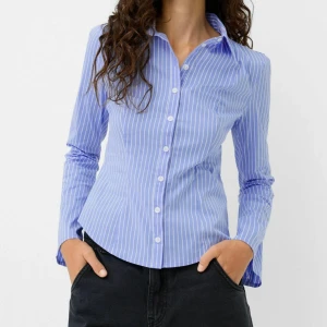 Blå- och vitrandig skjorta från Bershka - Snygg blå skjorta med vita ränder från Bershka. Skjortan har klassisk krage, långa ärmar och knäppning framtill. Oanvänd, endast testad med lappen kvar. 