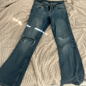 Low Waits Blå bootcut jeans - Säljer ett par blå bootcut jeans med hål på ena benet. Jeansen har klassisk femficksdesign och normal passform upptill. Perfekta för dig som gillar en avslappnad och trendig look.