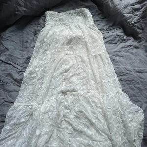 Vit långkjol från Gina Tricot - Säljer en vit långkjol från Gina Tricot med smock i midjan och diskret broderat mönster. Kjolen har flera volanger och är tillverkad i ett lätt tyg som ger en luftig känsla. Perfekt för varma dagar eller när du vill ha en romantisk look.