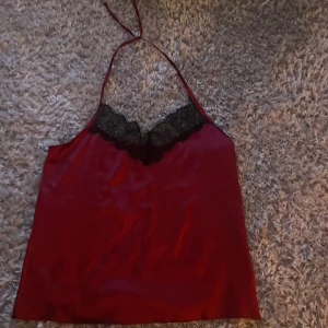 Röd satin linnetopp med spetsdetalj - Snygg röd linnetopp i satin med svarta spetsdetaljer vid bysten och tunna axelband som knyts i nacken. 