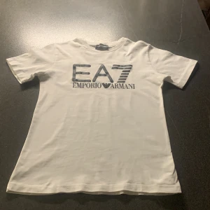 Vit t-shirt från Emporio Armani EA7 - Säljer en stilren vit t-shirt från Emporio Armani EA7 med svart logga tryckt på bröstet. T-shirten har klassisk passform och rund hals. Perfekt för dig som gillar exklusiva märken och enkel design.