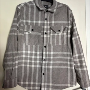 Grå och vit rutig overshirt från H&M - Säljer en grå och vit rutig overshirt från H&M. Skjortan har långa ärmar, två bröstfickor med knappar och klassisk krage. Perfekt att bära som lager över en t-shirt. Stängs med svarta knappar framtill.                        Använd fåtal gånger
