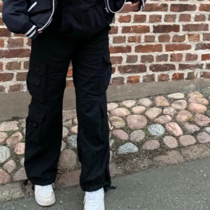 Svarta cargopants med fickor - Säljer ett par svarta cargopants med flera stora fickor på sidorna och avslappnad passform. Byxorna har dragsko vid benslut och är perfekta för en streetwear-look. Passar dig som gillar praktiska och trendiga byxor. 