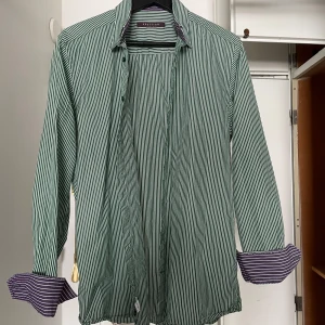 Grön och vit randig skjorta - Snygg grön och vit randig skjorta med klassisk krage och långa ärmar. Manschetterna har ett kontrasterande lila och vitt mönster på insidan. Perfekt för dig som gillar stilrena detaljer. Storlek M