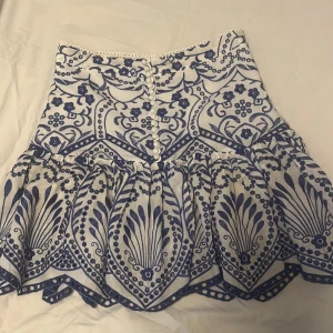 Vit och blå kjol med volang och mönster - Supersöt kjol i vitt och blått med dekorativa knappar framtill och volang nedtill. Kjolen har ett detaljerat blommigt och grafiskt mönster och är perfekt för dig som gillar unika plagg med mycket detaljer. Ingen lapp kvar men den är helt oanvänd!