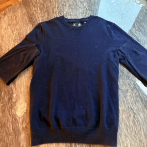 Mörkblå tröja från Lyle & Scott - Säljer en stilren mörkblå tröja från Lyle & Scott med rund halsringning och diskret broderad logga på bröstet. Tröjan har långa ärmar och är tillverkad i mjukt material som passar perfekt till vardags.