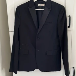 Svart kavaj från H&M - Stilren svart kavaj från H&M med klassisk passform och två knappar framtill. Kavajen har släta slag, tre fickor på framsidan och är helfodrad. Perfekt för formella tillfällen eller bal.