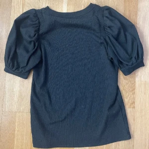 Svart topp med puffärmar - Säljer en svart topp med korta puffärmar och rund halsringning. Toppen har en lätt ribbad struktur och en klassisk, stilren look som passar till många outfits.