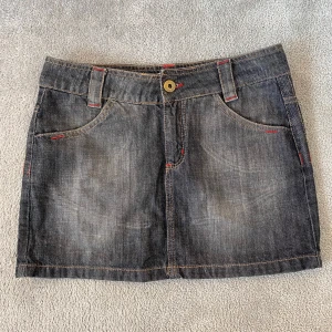 Y2k Jeanskjol från Miss P  - Mörkgrå jeanskjol med röda sömmar och klassiska fickor fram. Kjolen har bälteshällor och stängs med knapp och dragkedja. Perfekt för en cool och avslappnad stil.