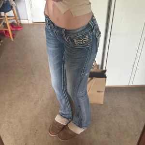 Blå jeansbyxor med broderade detaljer , Rock revival - Säljer ett par blå jeansbyxor med snygga broderade fickor och vita kontrastsömmar. Byxorna har låg midja och en bootcut passform. Perfekta för dig som gillar detaljer och en klassisk jeanslook. Köpta för 1300kr, kan mötas i stockholm eller frakta men står ej för frakten! Kontakta för mer frågor! (De är nu uppklippa längst nere för att få de längre!!)