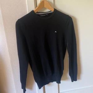 Svart stickad tröja från J.Lindeberg - Klassisk svart stickad tröja från J.Lindeberg med rund halsringning och diskret logga på bröstet. Perfekt basplagg med lång ärm och smal passform. Enkel och stilren design som passar till mycket.