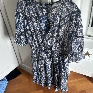 Blåvit mönstrad omlottklänning från Zara - Säljer en blåvit omlottklänning från Zara med blommigt mönster och trekvartsärm. Klänningen har v-ringning och resår i midjan för en smickrande passform. Perfekt för vår och sommar!