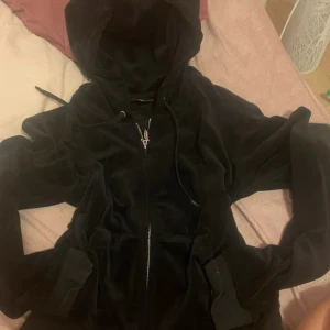 Svart velour hoodie från Juicy Couture - Mysig svart hoodie i velour från Juicy Couture med dragkedja framtill och huva. Klassisk modell med fickor på sidorna och justerbar dragsko i huvan. Perfekt för en avslappnad och trendig look.