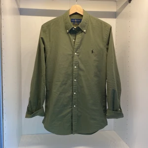 Olivgrön skjorta från Ralph Lauren - Klassisk olivgrön skjorta från Ralph Lauren med broderad logga på bröstet. Skjortan har knappar framtill och på kragen samt långa ärmar. Säljer den då den har blivit för liten, väldigt bra skick då den inte hunnit med mycket användning😃
