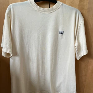Beige t-shirt med WB-logga - Säljer en stilren off white T-shirt med liten WB-logga broderad på bröstet. T-shirten har rund halsringning och korta ärmar. Perfekt för en clean och enkel look.
