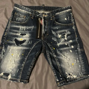 Jeansshorts från DSQUARED2 - Säljer ett par blå jeansshorts från DSQUARED2 med coola slitningar, färgstänk och en hjärtformad detalj. Shortsen har dragkedja framtill, klassiska fickor och DSQUARED2-logga på baksidan. Perfekta för dig som gillar streetwear och unika detaljer.