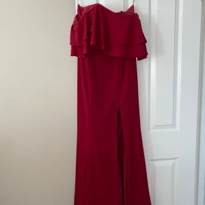 Rosaröd långklänning från Dressroom - Säljer en elegant röd långklänning från Dressroom med volang upptill och offshoulder-design. Klänningen har en slits framtill och är perfekt för speciella tillfällen. Materialet är mjukt och följsamt.