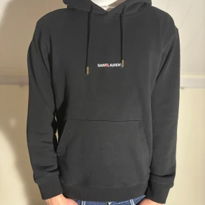 Svart hoodie från Saint Laurent - Säljer en svart hoodie från Saint Laurent med minimalistisk logga på bröstet. Tröjan har huva med dragsko och en stor magficka. Perfekt för dig som gillar stilrena och exklusiva plagg.