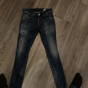 Mörkblå Dsquared2 jeans med slitningar - Säljer ett par mörkblå jeans från Dsquared2 med slitna detaljer och färgstänk. Jeansen har normal passform, klassisk femficksdesign och knappgylf. Märkeslogga syns vid gylfen. Perfekta för en trendig streetstil.