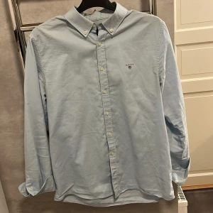 Ljusblå skjorta från Gant - Klassisk ljusblå skjorta från Gant med button-down krage och broderad logga på bröstet. Skjortan har långa ärmar och knäppning framtill. Perfekt för en stilren och avslappnad look.