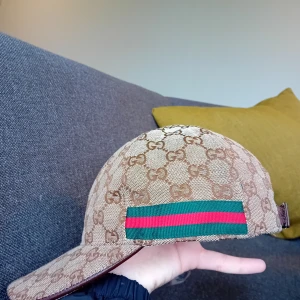 Gucci keps - Säljer en beige keps från Gucci med klassiskt GG-mönster över hela kepsen. Kepsen har en grön och röd randig detalj på sidan och justerbar rem i brunt läder baktill. Perfekt för dig som gillar exklusiva accessoarer.