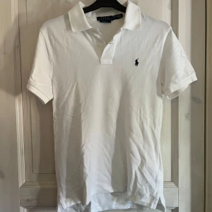 Vit pikétröja från Polo Ralph Lauren - Klassisk vit pikétröja från Polo Ralph Lauren med korta ärmar och krage. Tröjan har knappar vid halsen och den ikoniska broderade loggan på bröstet. Perfekt för en stilren och tidlös look.