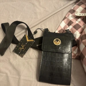 Svart axelväska med krokodilpräglat mönster från Vanzetti - Säljer en svart axelväska från Vanzetti med krokodilpräglat mönster och guldfärgad logga på locket. Väskan har justerbart axelband och stängs med lock. Perfekt för mobilen och småsaker.