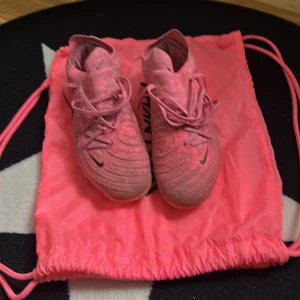 Nike Phantom GT fotbollsskor i rosa - Säljer ett par Nike Phantom GT fotbollsskor i en snygg rosa färg med svarta detaljer. Skorna har snörning och är tillverkade i syntetmaterial. Perfekta för dig som vill sticka ut på planen! Levereras med matchande rosa gympåse.